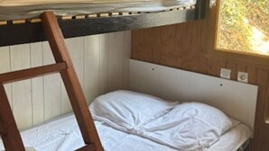 Una cama litera con escalera de madera, una ventana con vistas a follaje verde y una pared blanca con un reloj pequeño.
