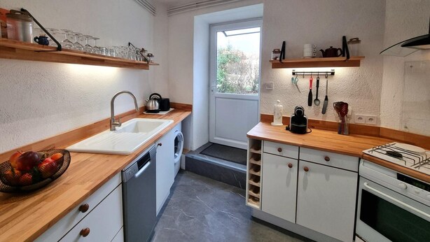 A compact kitchen with a dishwasher, sink, and a stove. There are wooden shelves above the counter and a window providing natural light. - Cozy village house - 4 guests (Bellegarde-en-Forez)