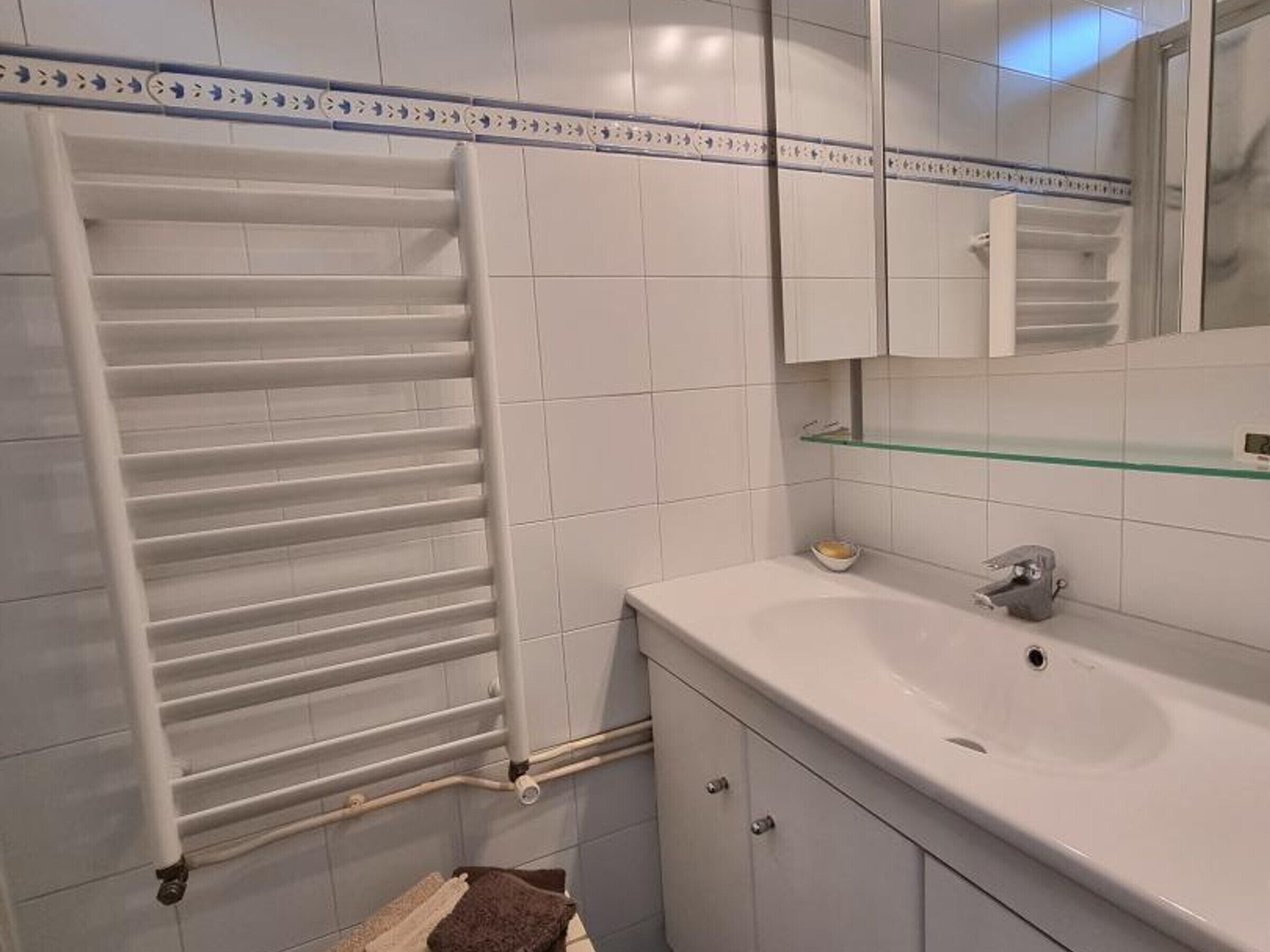 A bathroom with a white tiled wall, a glass shelf above the sink, a towel rack, and a cabinet with a mirror.
