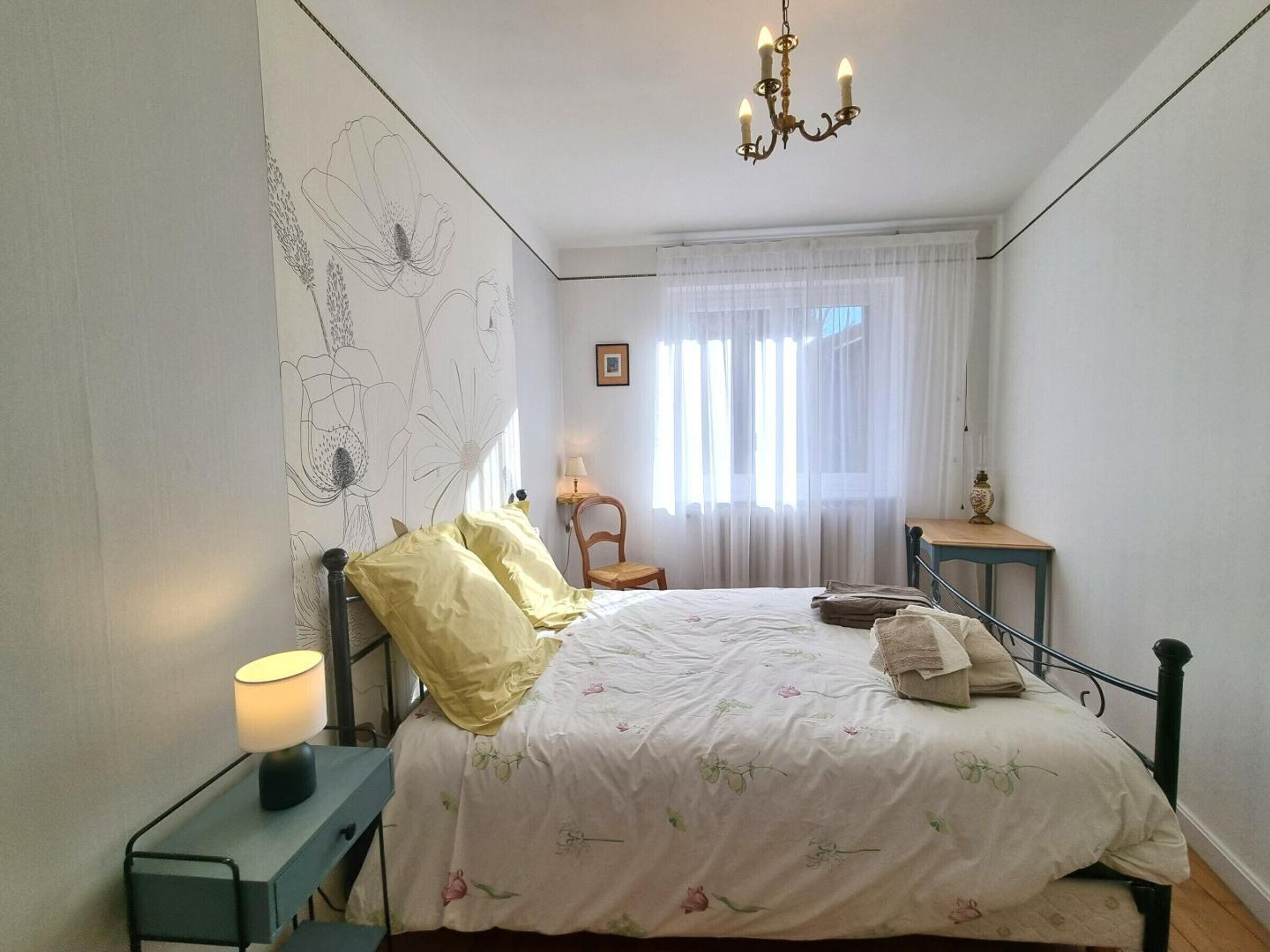 Ein Schlafzimmer mit Bett, Nachttisch, Stuhl, Tisch, einer Chandelier und einem Fenster mit Vorhängen.