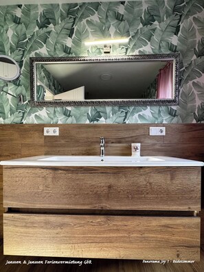 Une salle de bain avec un meuble-lavabo en bois, un grand miroir et un mur orné d’un motif végétal.