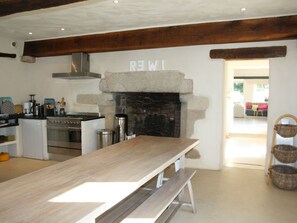 Une cuisine avec une cheminée en pierre, des poutres apparentes et une longue table en bois.