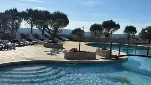 Zona de piscina con sillones de descanso y macetas, rodeada de árboles y un cielo despejado.