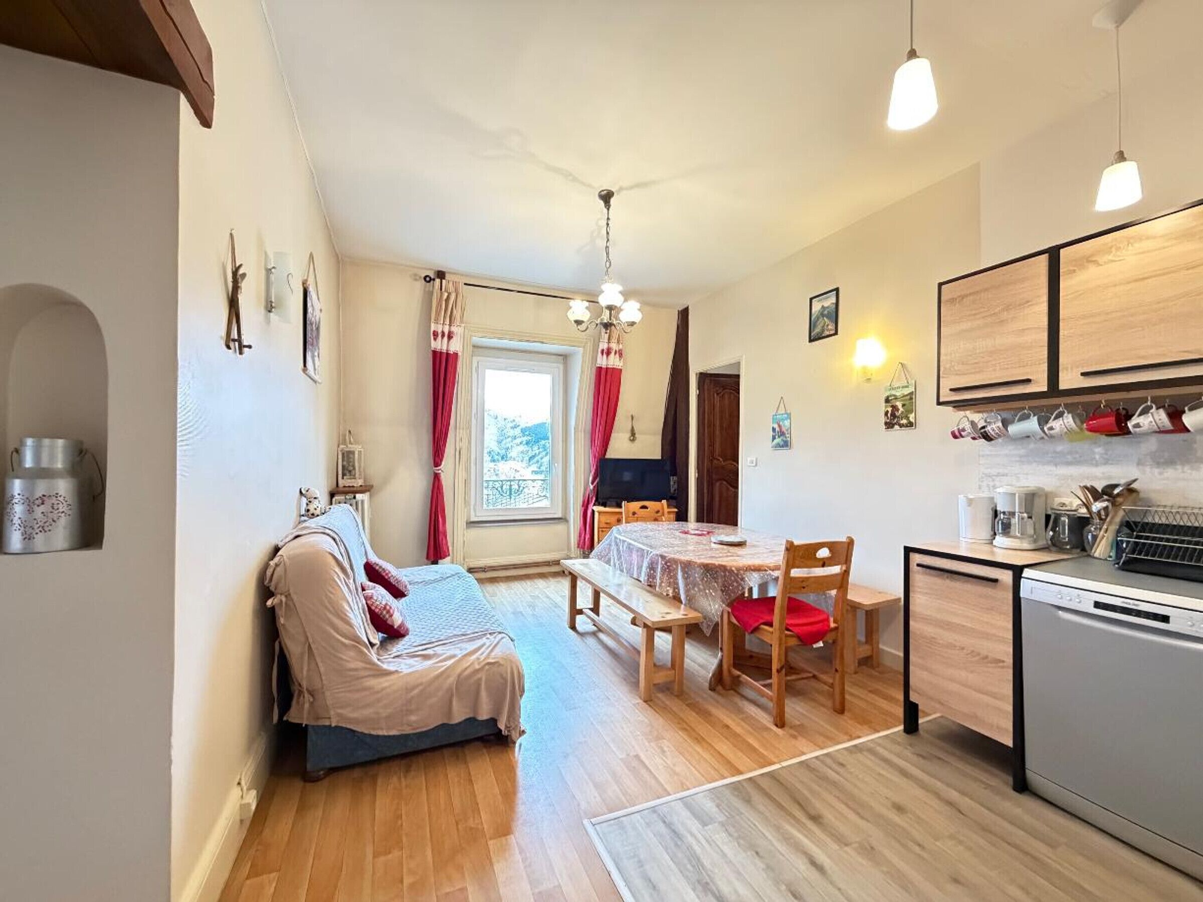 A cozy room with a bed, a dining table, chairs, a kitchen area with cabinets and a dishwasher, and a window with curtains.