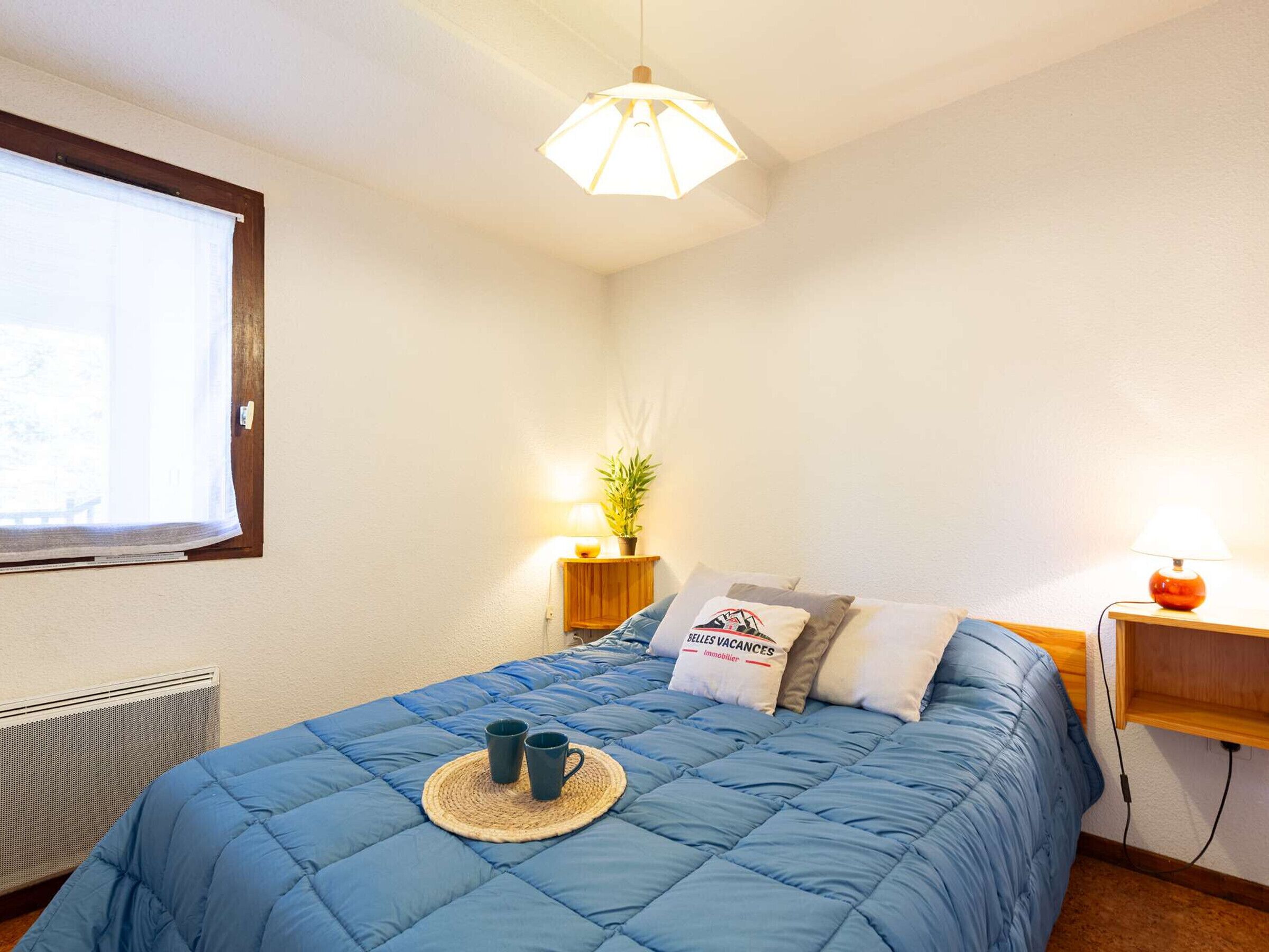 A bedroom with a blue bedspread, a bedside table with a lamp, a small plant, and a window with blinds.