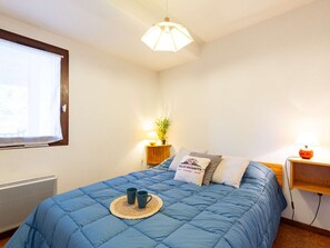 A bedroom with a blue bedspread, a bedside table with a lamp, a small plant, and a window with blinds. - Functional T3 with terrace and parking, Saint-Lary-Soulan (Saint-Lary-Soulan)