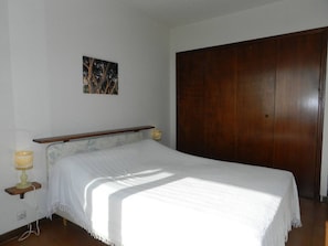 A bedroom with a bed, a wooden wardrobe, a nightstand with a lamp, and a wall-mounted picture. - Apartment sea view with parking - 4 guests (Bormes-les-Mimosas)