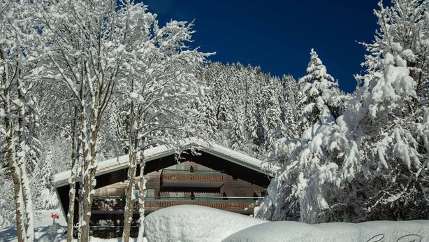 Un chalet enneigé, avec une façade en bois, entouré d’arbres chargés de neige.