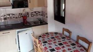 Une cuisine compacte équipée d’un four à micro-ondes, d’une machine à café et d’un petit réfrigérateur. On y trouve une table en bois recouverte d’une nappe à motifs, ainsi que deux chaises.