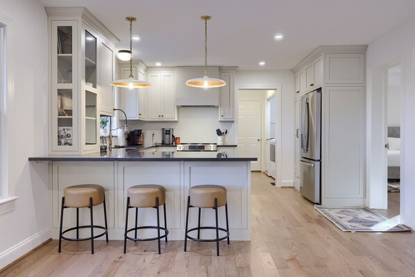 Una cocina moderna con armarios blancos, una isla central con taburetes, electrodomésticos de acero inoxidable e iluminación colgante.