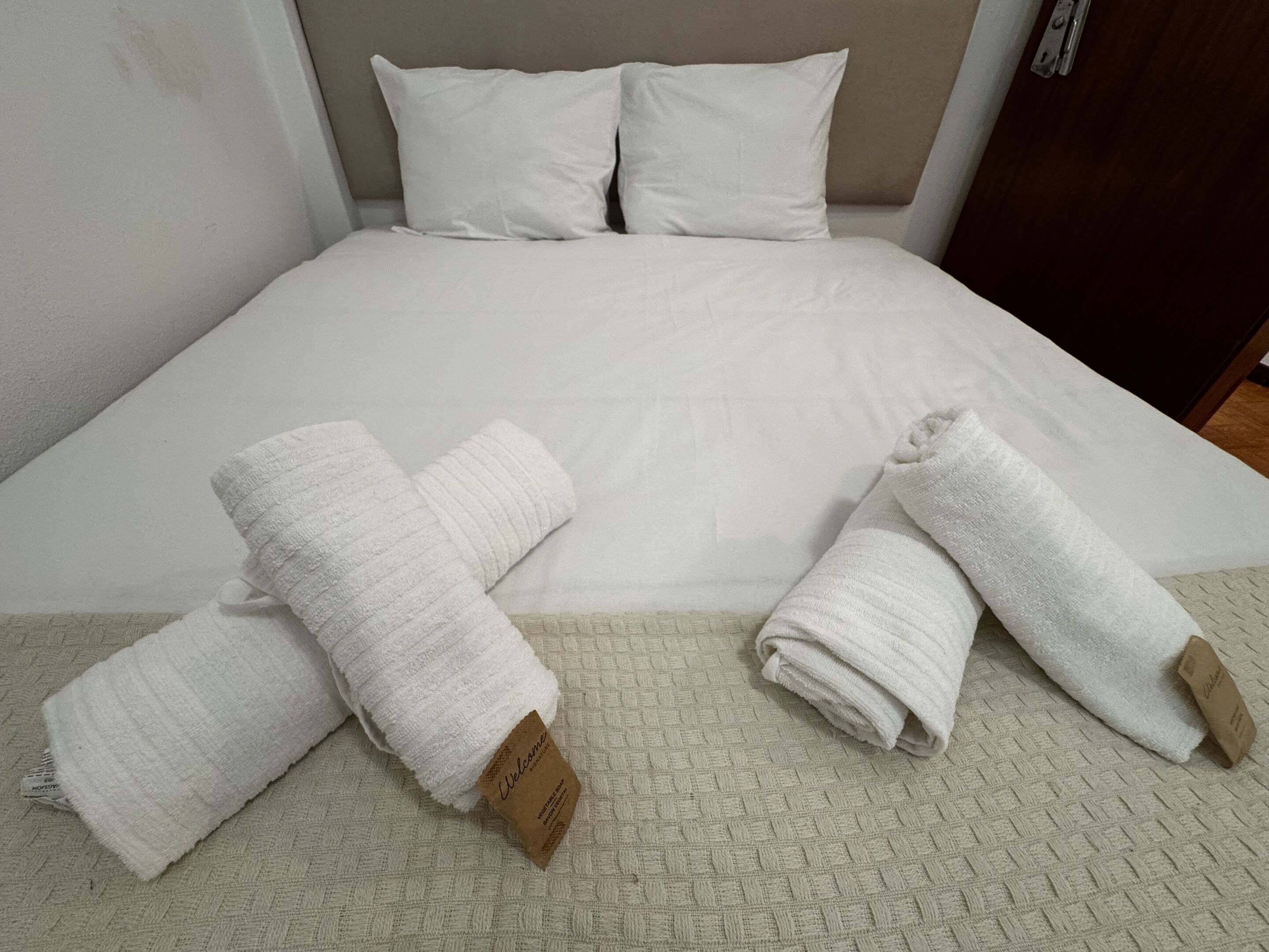 A bed with white bedding, two rolled towels with tags, and a textured floor.