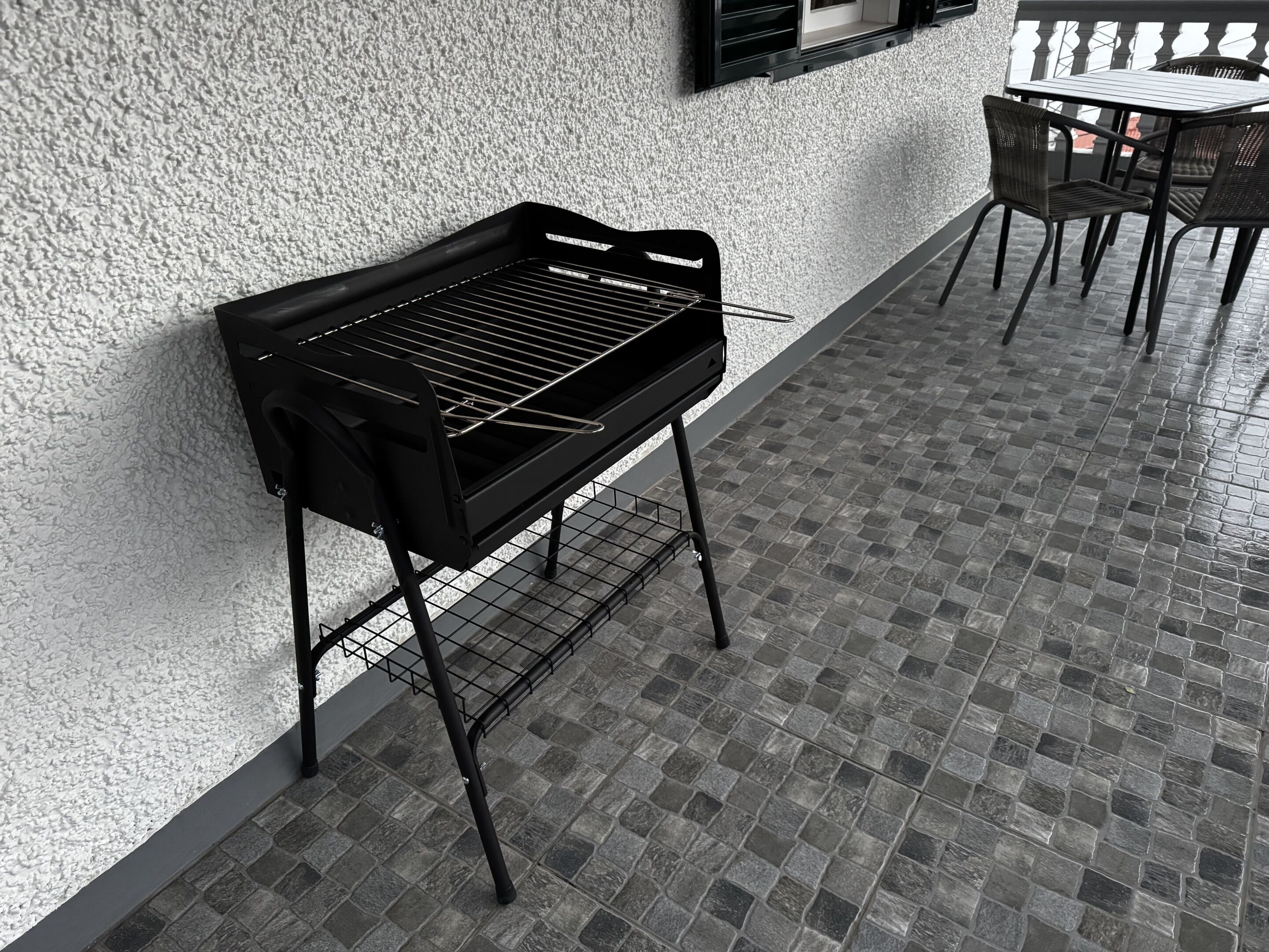 Una griglia per barbecue di fronte a una parete strutturata, un pavimento piastrellato e un tavolo con sedie sullo sfondo.