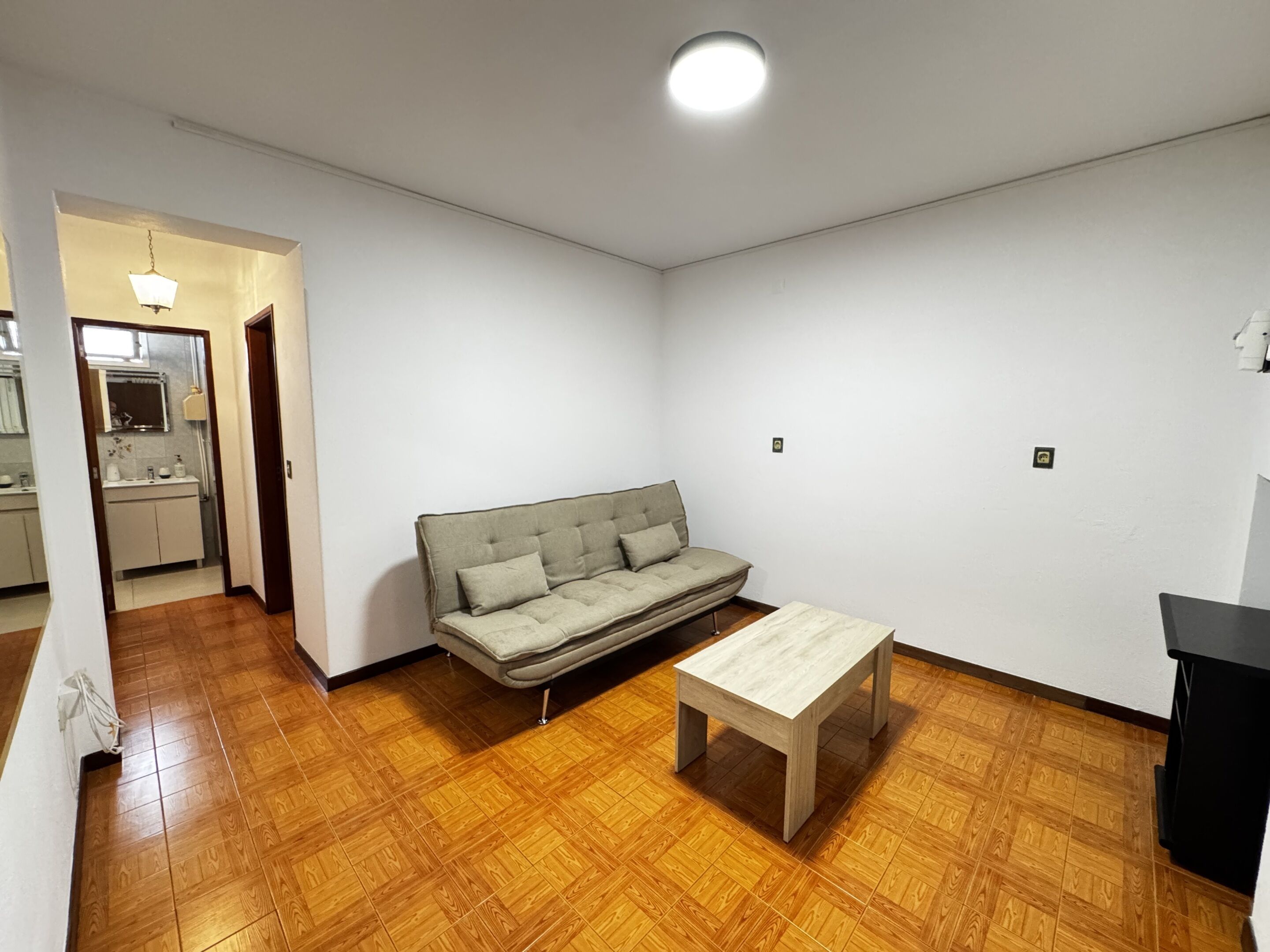 A room with a sofa, a small table, wooden flooring, and a kitchen area visible through an open doorway.