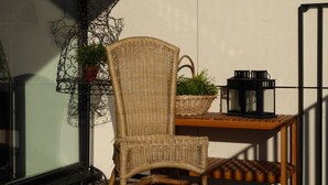 Een rieten stoel, een tafeltje met een lantaarn, en een plant op een balkon.