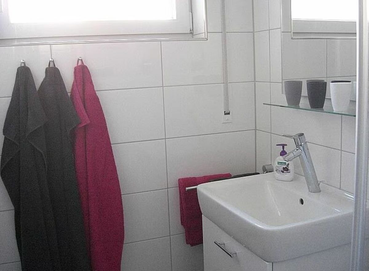 Een badkamer met witte tegels, een witte wastafel, een spiegel en drie handdoeken (zwart, roze en rood) die aan een rek hangen.