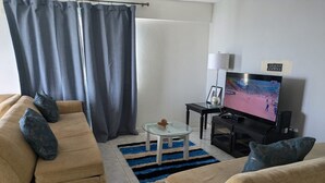Olohuoneessa on beige sohva, lasinen sohvapöytä, televisio ja sininen matto.