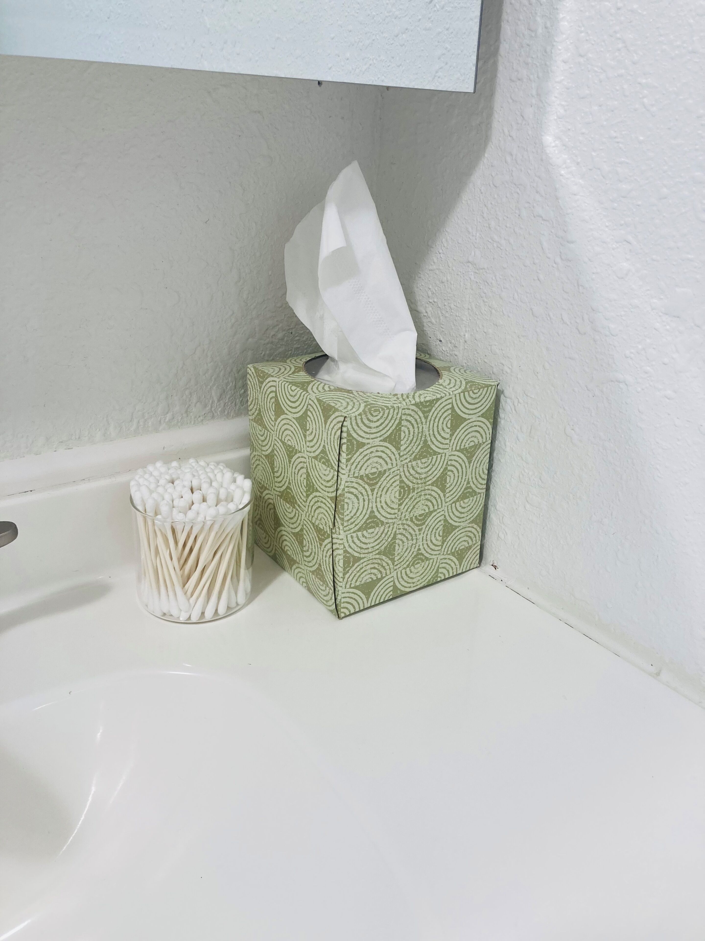 A tissue box with a green pattern and a container of cotton swabs on a white surface.