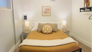 Un dormitorio ordenado con una cama, dos lámparas de noche, un aire acondicionado de pared, una obra de arte floral enmarco y un pequeño estante con objetos decorativos.