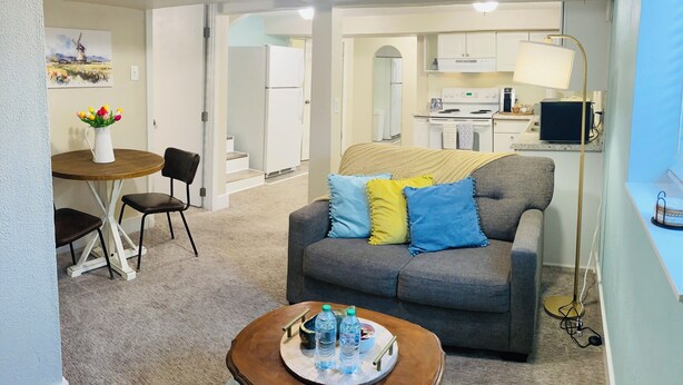 Un espacio habitable compacto con un sofá gris, una pequeña mesa de madera con una bandeja, una zona de comedor con una mesa y sillas blancas, y una kitchenette al fondo.