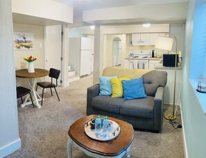 Un espace de vie compact comprenant un canapé gris, une petite table en bois avec un plateau, un coin repas avec une table et des chaises blanches, et une petite cuisine en arrière-plan.