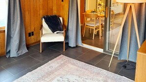 A room with wooden panel walls, a white chair with a black cushion, a patterned rug, a wooden floor, and a large mirror reflecting a dining area. - Summer Retreat in Traverse City: Steps to TART Trail & State Park Beach (Traverse City)