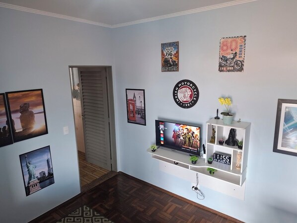 A living room with a TV, wall art, a clock, and a shelf with plants.