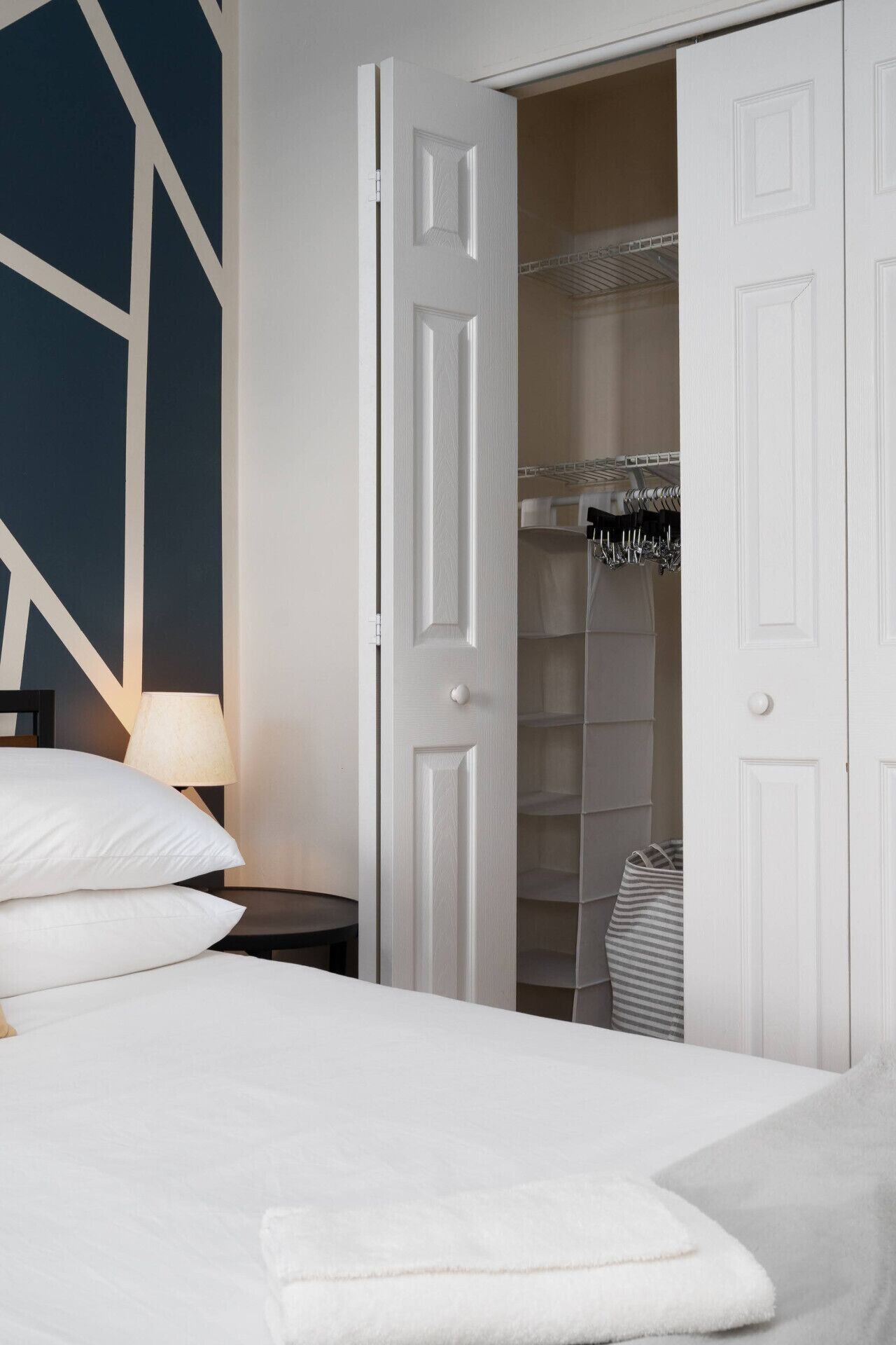 Une chambre à coucher avec un lit, une table de chevet, une lampe, une armoire où sont suspendus des vêtements, et un sac à rayures.