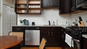A kitchen with dark cabinets, a stainless steel dishwasher, a gas stove, and a dining area with a wooden table and chairs. - Flexhome Ukrainian Village • 2BR • Walkable + Private Balcony (Chicago)