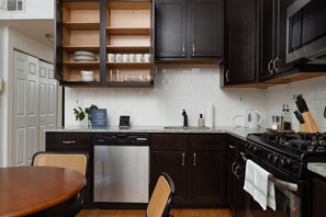 A kitchen with dark cabinets, a stainless steel dishwasher, a gas stove, and a dining area with a wooden table and chairs. - Flexhome Ukrainian Village • 2BR • Walkable + Private Balcony (Chicago)