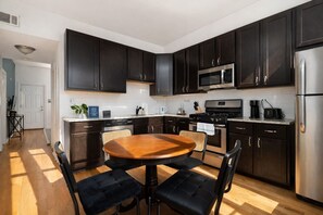 A modern kitchen with dark cabinets, stainless steel appliances, a wooden dining table, and black upholstered chairs.