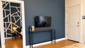 A room with a wooden floor, a TV mounted on the wall, a small table with books, and a door with a 'No Smoking' sign. - Flexhome Ukrainian Village • 2BR • Walkable + Private Balcony (Chicago)