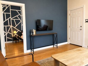 A room with a wooden floor, a TV mounted on the wall, a small table with books, and a door with a 'No Smoking' sign. - Flexhome Ukrainian Village • 2BR • Walkable + Private Balcony (Chicago)