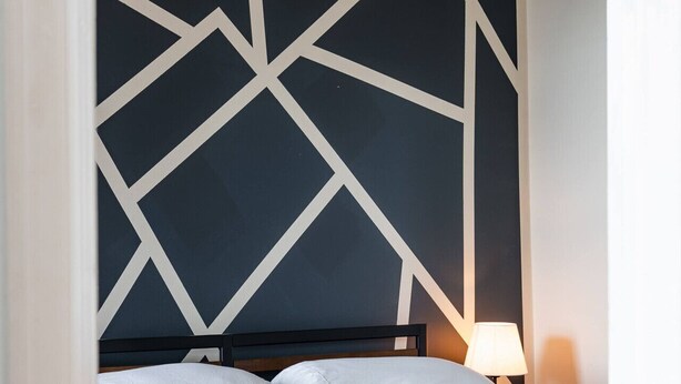 A neatly made bed with white linens and a grey throw blanket, a bedside lamp, and a geometric wall design. - Flexhome Ukrainian Village • 2BR • Walkable + Private Balcony (Chicago)