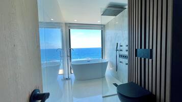 A modern bathroom with a bathtub, a toilet, and a shower area, featuring a large window with a view of the ocean.
