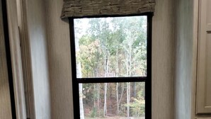 Ein Fenster mit Blick auf einen Wald, eine Sitzbank und ein Bett mit gemustertem Überwurf.