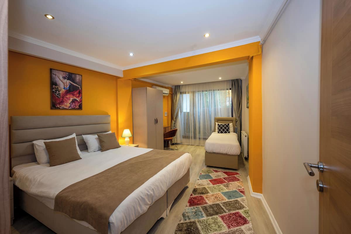 A hotel room with a large bed, a smaller bed, a desk, and a chair. There is a painting on the wall and a window with curtains.