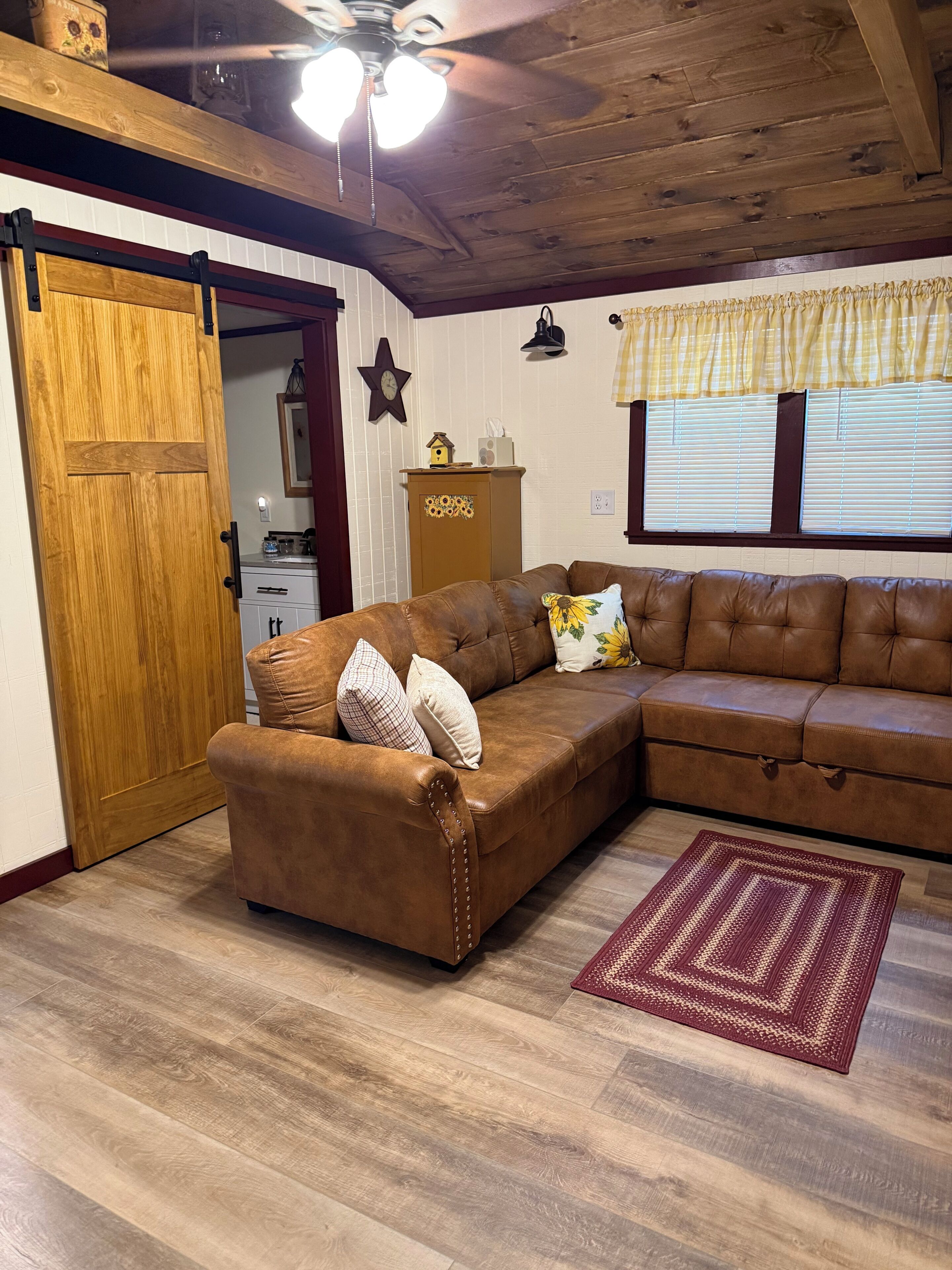 A cozy living room with a brown leather sectional sofa, a wooden door, a star-shaped wall decoration, a small cabinet, and a window with blinds.
