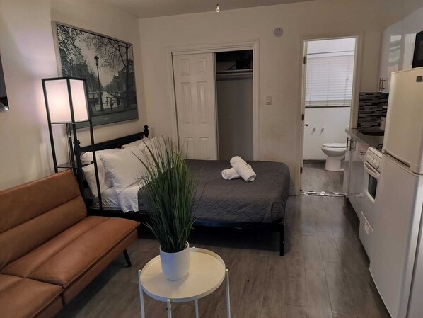 A compact hotel room with a bed, a small table with a plant, a sofa, a painting on the wall, a closet, and a kitchenette. - Comfort & Convenience Right Here F (Hollywood)