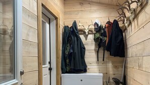 Un couloir étroit aux murs en bois, une porte blanche et un appareil électroménager blanc. Des bancs et des bottes sont disposés le long des murs, et des vêtements sont suspendus à des crochets.