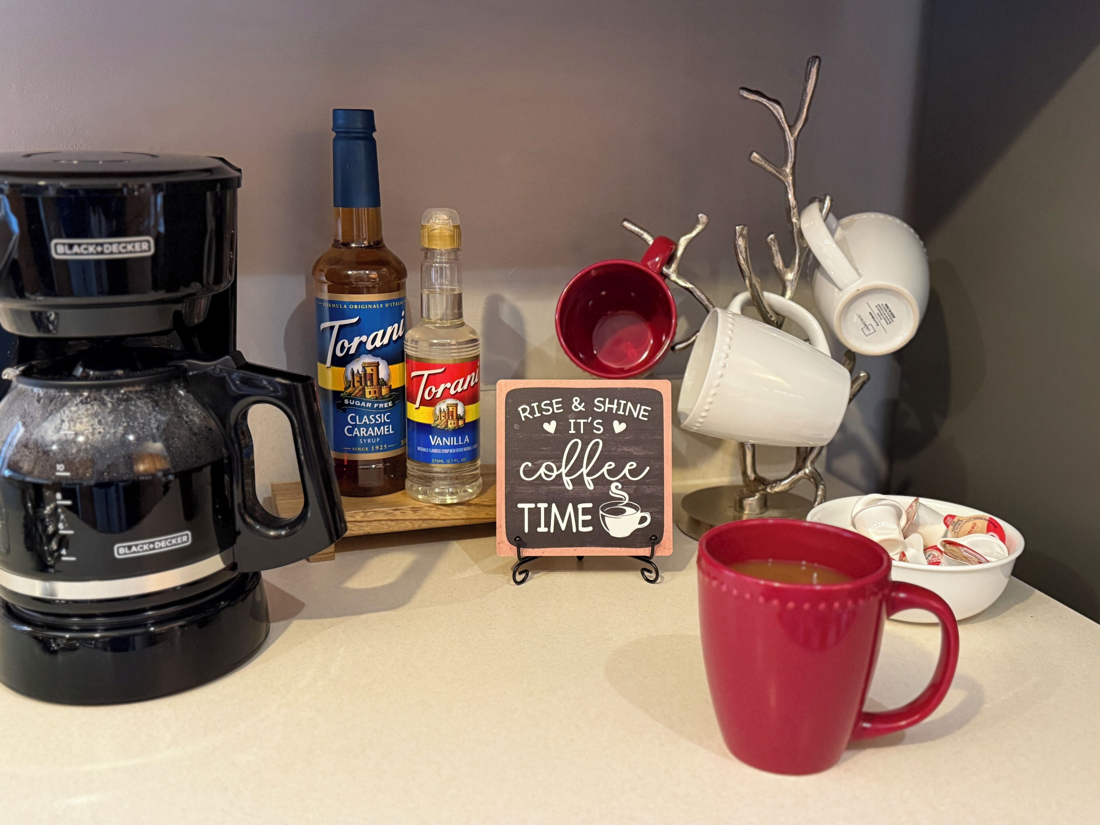 A coffee maker, Torani syrup bottles, a Torani vanilla bottle, a red mug, a coffee-themed sign, and white mugs on a countertop.
