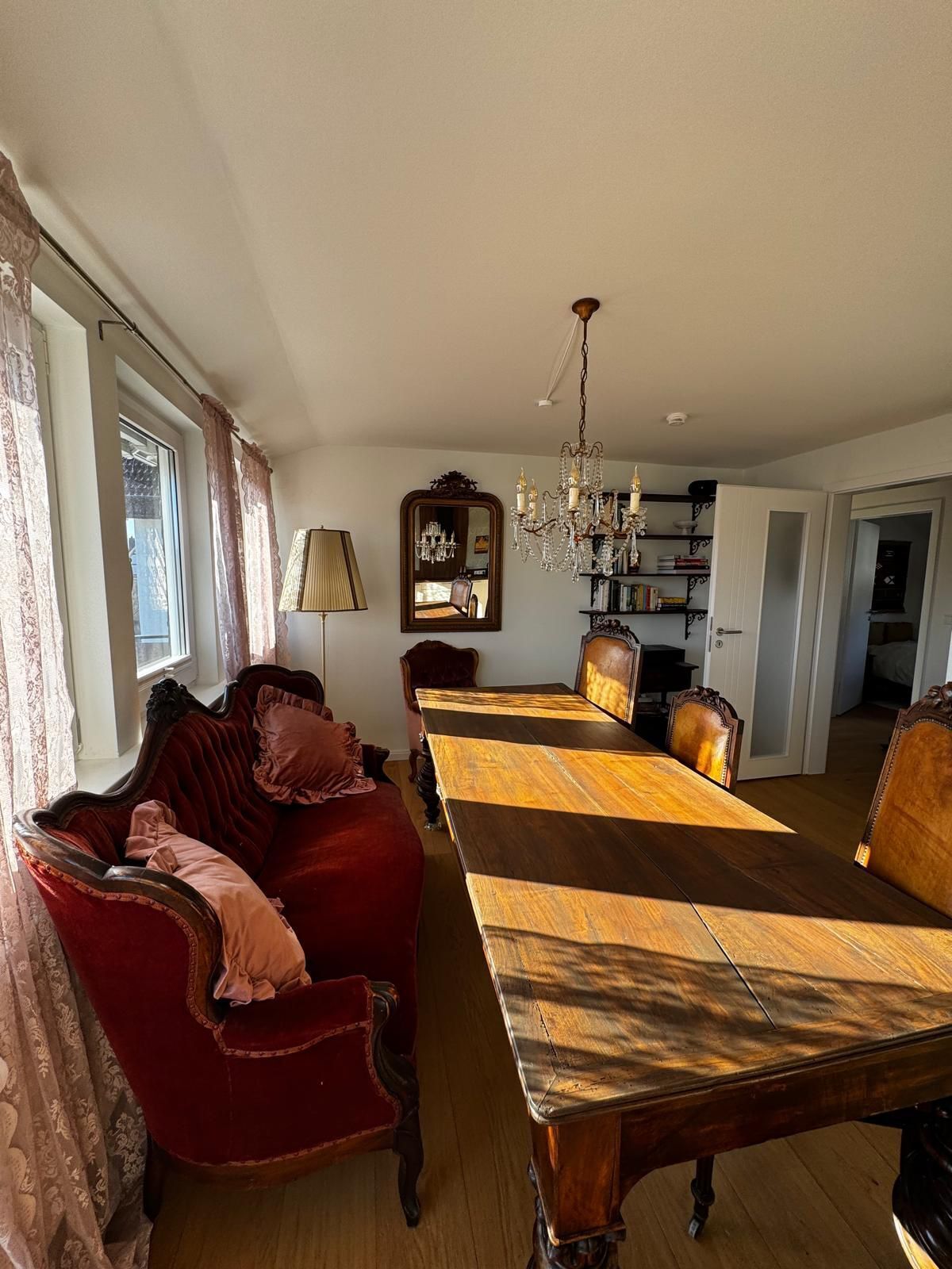 Une salle à manger avec une longue table en bois, un canapé rouge recouvert de tissu, un lustre et un miroir.