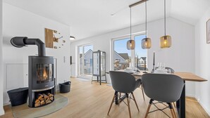 A modern kitchen with a wood-burning stove, a dining area with a table and chairs, and a view of houses through large windows. - Havhus - Sauna, fireplace, washing machine, electric vehicle charging station (Olpenitz)