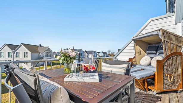 A wooden deck with a table, wicker furniture, and a view of houses in the background. - Havhus - Sauna, fireplace, washing machine, electric vehicle charging station (Olpenitz)