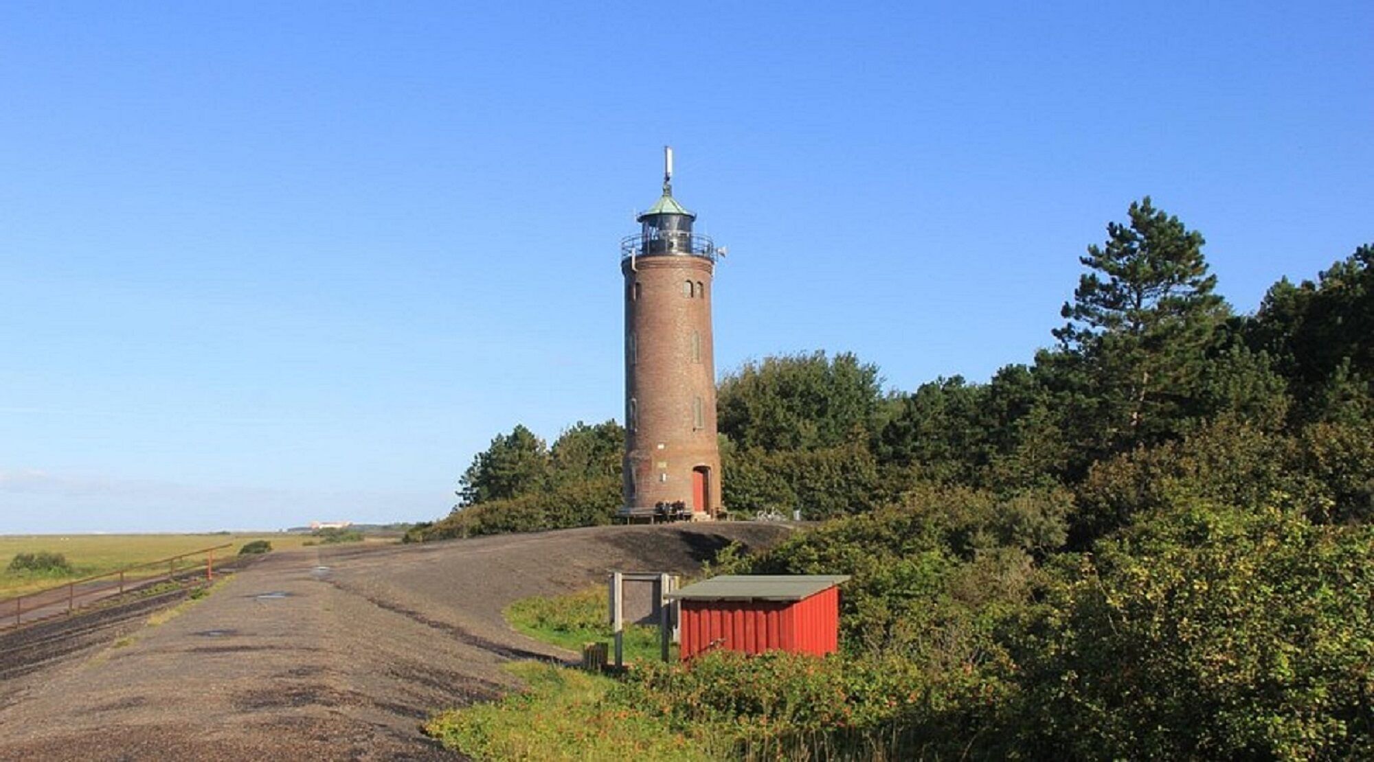 Ein Leuchtturm mit Backsteingrundlage und Metallkuppel, umgeben von Grünpflanzen und einem Kiesweg.