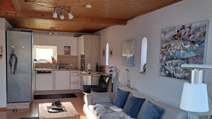 Una sala de estar moderna con un sofá, una mesa de centro y una lámpara. Se ve una cocina con armarios y un refrigerador al fondo.