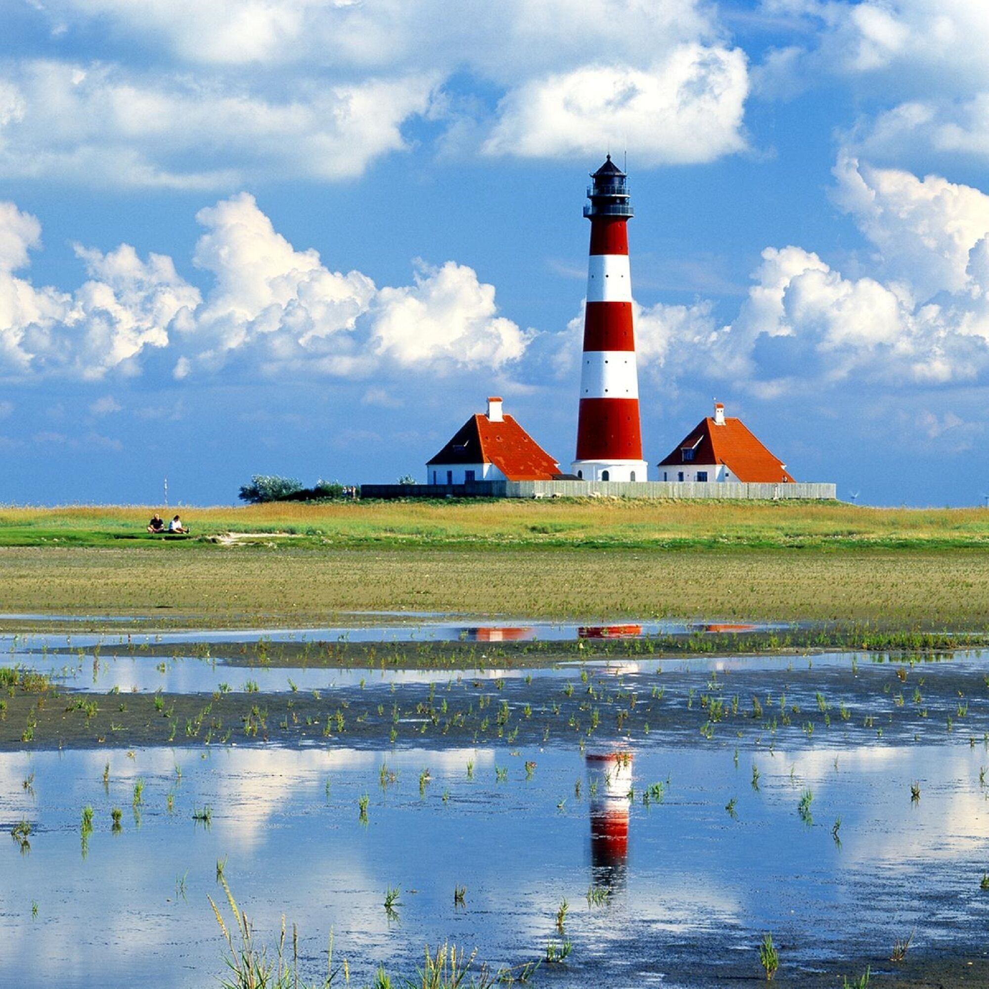Un phare doté d’une tour rayée de rouge et de blanc, deux bâtiments blancs avec des toits rouges, et une étendue d’eau calme qui reflète les constructions.