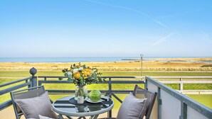 En balkong med ett bord, två stolar, en vas med blommor, en vattenmelon och utsikt över stranden och havet.