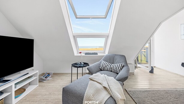 Ett modernt sovrum i loftstil med ett takfönster, en tv, en grå soffa och utsikt över omgivningen.