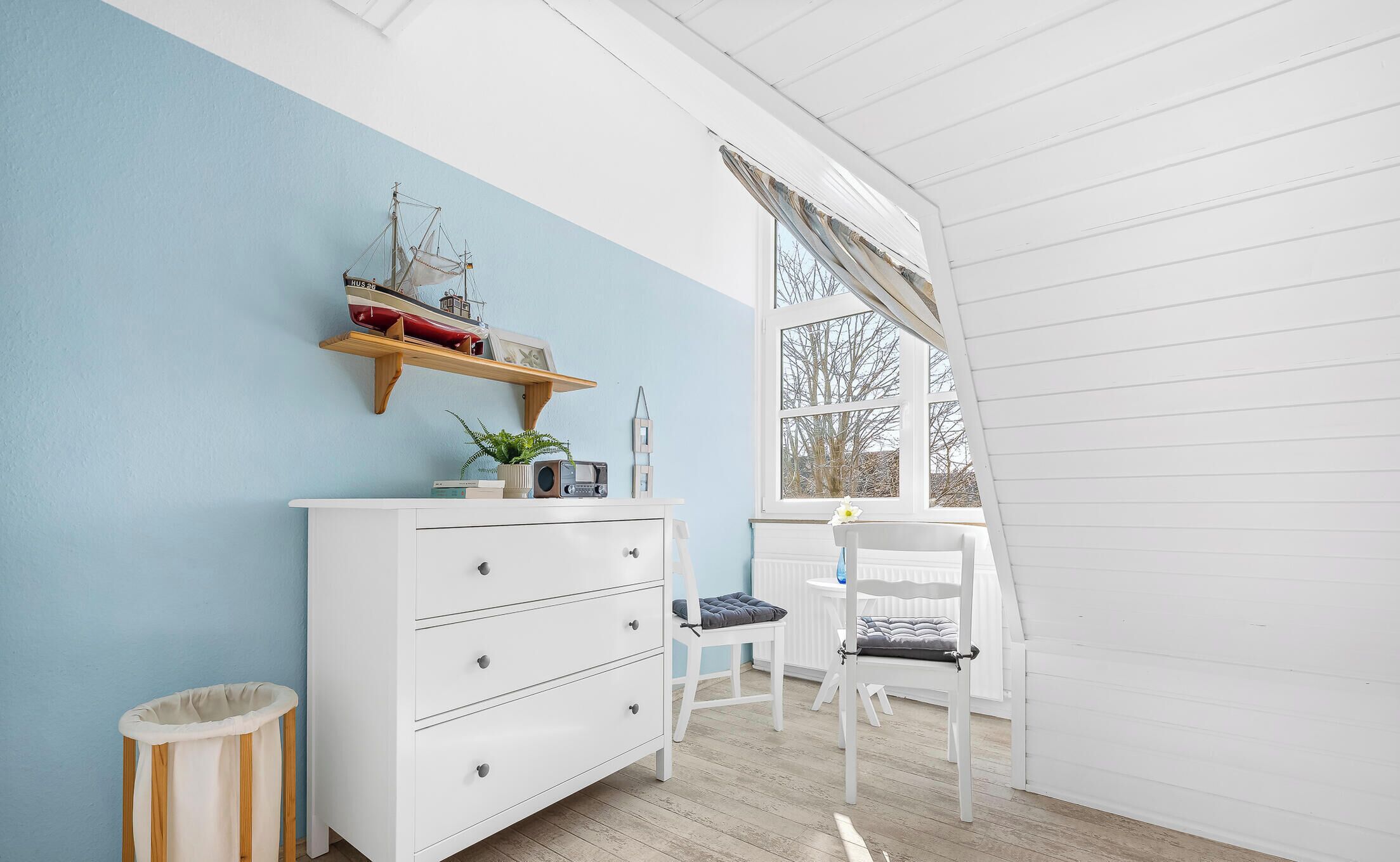 A room with a white dresser, a chair, a window with a view, and a shelf with decorative items.