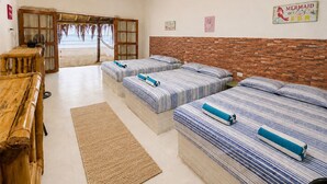 Hotellihuone, jossa on kolme sänkyä, puinen penkki, näkymä rannalle ja tiiliseinä.